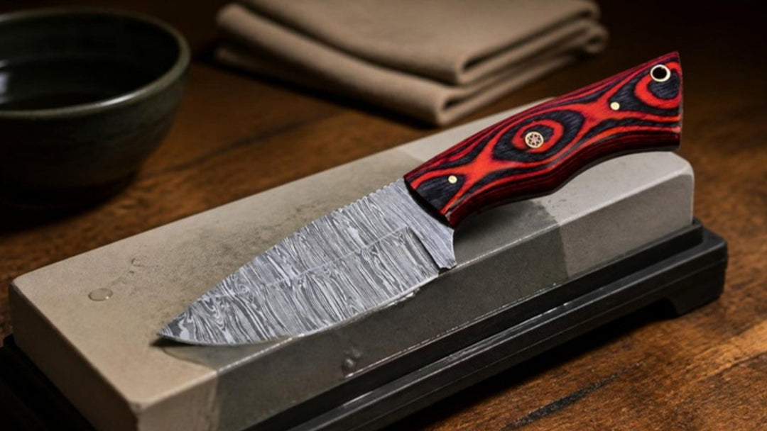 Damascus hunting knife with red and black Pakka wood handle resting on a wet whetstone